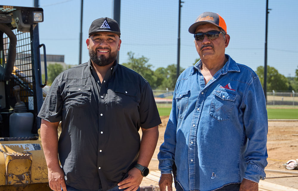 Two men, Benny and Leno Aguilar, stand side by side at a construction site in front of heavy machinery.
